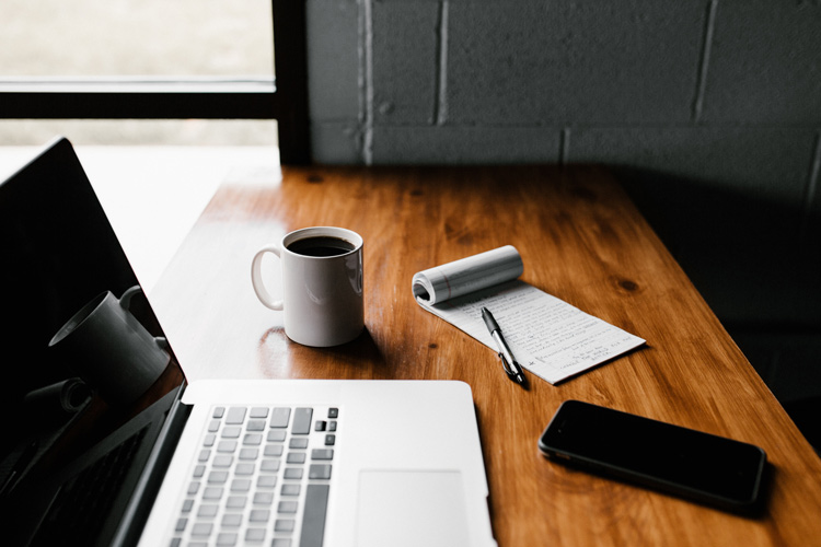 A laptop, mug, notepad, pen, and mobile phone on a wood desk